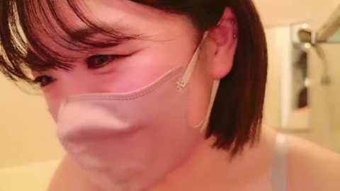 Media: Video of an Asian woman with short black hair and light skin, wearing a white surgical mask, in a bathroom with a glass shower door and a towel rack.