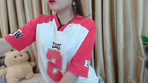 Media: Video of a young woman with light skin, wearing a red and white football jersey with number 2, holding a teddy bear, against beige curtains.