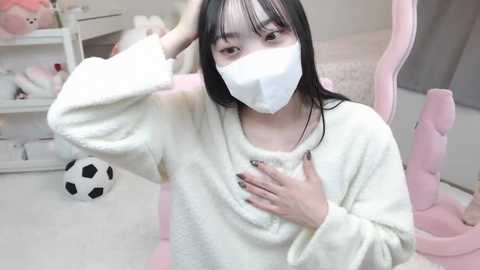 Media: Video of an East Asian woman with long black hair, wearing a white mask, fluffy white sweater, and pink chair, in a pastel-colored bedroom with toys and a soccer ball.