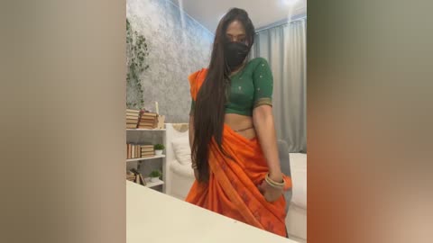 Media: Video of a South Asian woman in a green top, orange sari, and black face mask, standing in a modern room with white walls, shelves, and a bed.