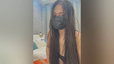 Media: Video of a woman with long, straight black hair, wearing a black mask and black bra, standing in a modern bedroom with a white bed, blue curtains, and a gold lamp.