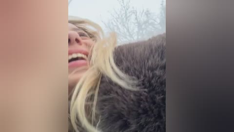 Media: Video of a smiling, fair-skinned woman with blonde hair partially covered by a dark brown fur coat, taken in a snowy outdoor setting with bare trees in the background.