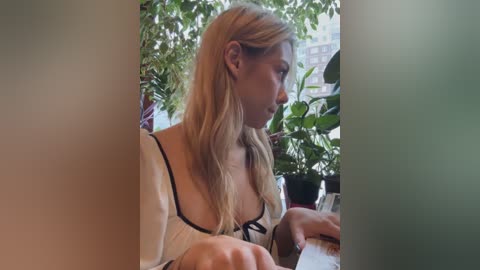 Media: A video of a blonde woman with long hair, wearing a white blouse, sitting inside a window frame, surrounded by green plants, focused on reading a book.