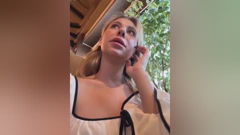 Media: Video of a fair-skinned woman with blonde hair, wearing a white blouse with black trim, talking on a phone outdoors amidst greenery.