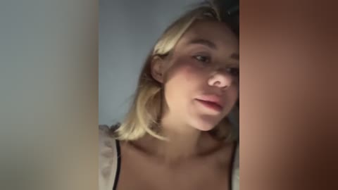 Media: Video of a young, light-skinned woman with shoulder-length blonde hair, wearing a white blouse, standing in a dimly lit room with a brown door partially blocking the view.