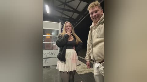 Media: Video of a blonde woman with long hair in a black jacket, pink skirt, and black tights, standing with a man in a beige hoodie and light pants, both smiling near a graffiti-covered bus stop.