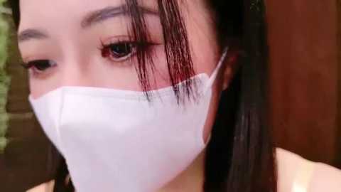 Media: Video of an Asian woman with fair skin, dark hair, and long bangs partially covering her face. She wears a white surgical mask. Background shows a blurred wooden door and green foliage.