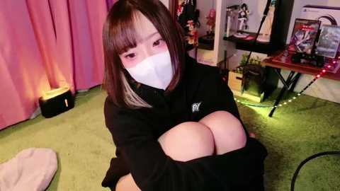 Media: Video of an Asian woman with straight brown hair, wearing a black sweater, white face mask, and a white bra exposing large breasts. She sits on a green carpeted floor in a dimly lit room with pink curtains, gaming equipment, and Christmas decorations.