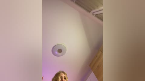 Media: Video of a blonde woman with shoulder-length hair, partially visible, standing in a dimly lit room with white walls and a circular ceiling light. The room has a minimalist, modern aesthetic.