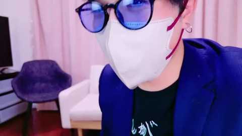 Media: Video of a person wearing a white mask, blue glasses, and a purple jacket, sitting in a room with a white chair, purple chair, and pink curtains in the background.