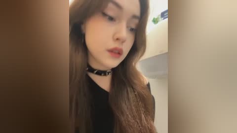 Video of a young, fair-skinned woman with long brown hair, wearing a black choker and a black top, gazing downwards in a dimly lit room with beige walls.