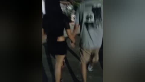 Media: Video of a blurred, dark-skinned woman in a black top and shorts, walking past a blurred white man in a grey shirt. Background is dimly lit, possibly an urban setting.