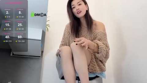 A video of an East Asian woman with long black hair, wearing a knitted off-shoulder sweater and white shorts, seated in a modern, minimalist room.