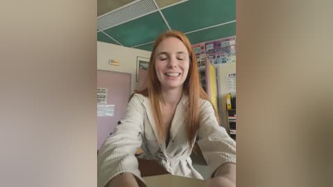 Media: Video of a smiling, fair-skinned, red-haired woman with long hair, wearing a white button-down shirt, sitting in a classroom with green ceiling tiles, posters, and bulletin boards in the background.