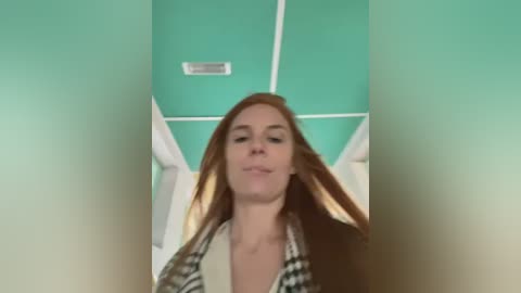 Media: A video of a young woman with long red hair, wearing a black jacket and plaid shirt, standing in a teal-painted room with white trim and a vent on the ceiling.