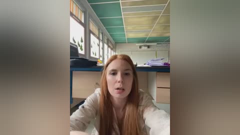 Media: Video of a young Caucasian woman with long, straight red hair, wearing a beige sweater, leaning over a desk in a classroom. Background features green and yellow ceiling tiles, windows with colored glass, and a cluttered desk.