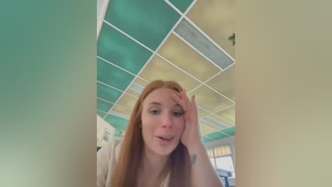 Media: Video of a young red-haired woman with fair skin, wearing a white shirt, sitting in a room with a teal and yellow geometric ceiling and large windows. She appears to be resting her head on her hand.