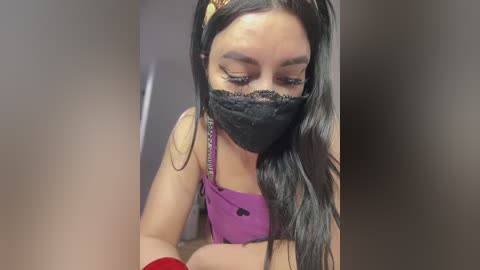 Media: Video of a young woman with long black hair, wearing a black face mask, purple sleeveless top, and red wristbands, looking down. Background is blurred.