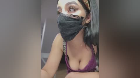 Media: Video of a woman with dark hair, wearing a black face mask, revealing a cleavage in a low-cut purple lace bra, leaning forward in a dimly lit room.