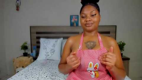 Media: Video of a smiling, dark-skinned woman with braided hair, wearing a red and white checkered apron, thumbs-up, in a modern bedroom with white bedspread and minimal decor.