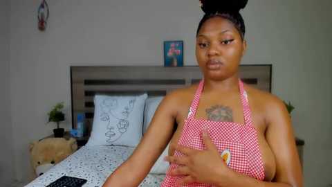 Media: Video of a young Black woman with a medium complexion and braided hair, wearing a red gingham apron, standing in a bedroom with a white bed and a wooden headboard.