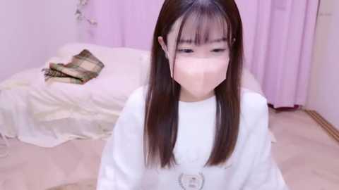 Media: Video of a young Asian woman with straight, long black hair, wearing a white mask, white sweater, and a black necklace, standing in a pastel-colored, softly lit room with a plaid blanket on a couch.