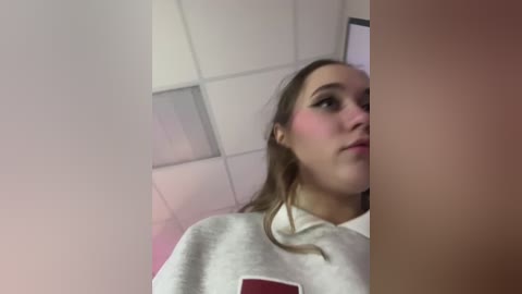 Media: Video of a young woman with light skin, brown hair, and light makeup, wearing a white sweatshirt, standing in a white-tiled room with fluorescent lights.