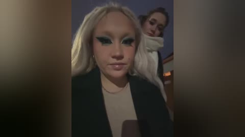 Media: Video of two young women: blonde in foreground, wearing dark jacket, green eyeshadow; brunette in background, wearing white sweater, looking distressed.