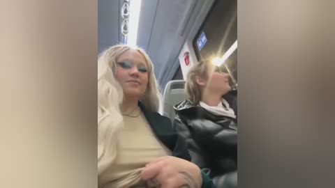 Media: Video of two women seated in an airplane. One woman with platinum blonde hair and blue eyeshadow, wearing a beige sweater, looks relaxed. The other, with light brown hair, wears a black jacket.