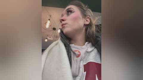 Media: Video of a young woman with fair skin and light brown hair in a ponytail, wearing a white and red sweatshirt. She has a neutral expression, looking upwards. Background features a modern ceiling with pendant lights.