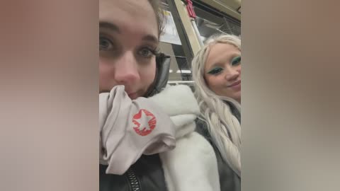 Media: Video of two women in a subway car. One, with long blonde hair and green eyeshadow, smiles. The other, with brown hair, has a white beanie with a red logo.