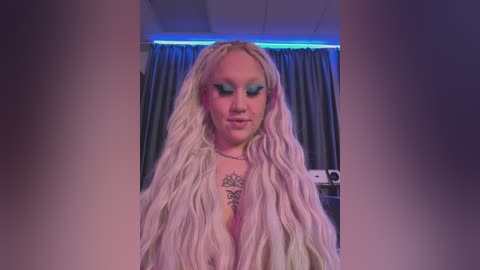 Media: Video of a young woman with long, wavy, platinum blonde hair, wearing a black top, and vibrant turquoise eye makeup, standing in a dimly lit room with blue lighting and gray curtains.