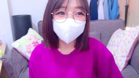 Media: Video of an Asian woman with straight, shoulder-length brown hair, wearing a white face mask, round glasses, and a bright pink top. She's seated on a gray sofa adorned with floral-patterned cushions in a cozy, indoor setting.