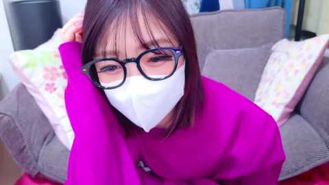 Media: Video of an East Asian woman with straight, shoulder-length brown hair, wearing black-rimmed glasses, a white face mask, and a bright pink sweater. She sits on a grey couch with floral-patterned cushions, indoors.