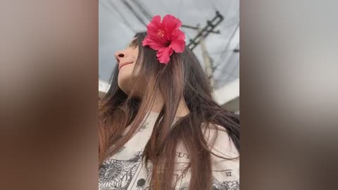 Media: Video of a young woman with long, dark hair, wearing a floral headband with a bright pink flower. She has a soft, gentle smile, and is dressed in a white, patterned shirt. The background is blurred, featuring a white ceiling with exposed beams.