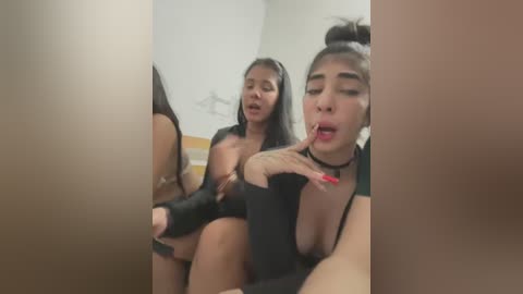 Media: Video of two women, one Asian with dark hair, wearing a black top, and the other with long hair, licking a finger, in a bathroom with beige tiles.