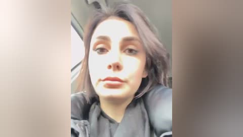 Media: Video of a young woman with light skin and dark hair, wearing a black jacket, seated in a car with a blurred background.