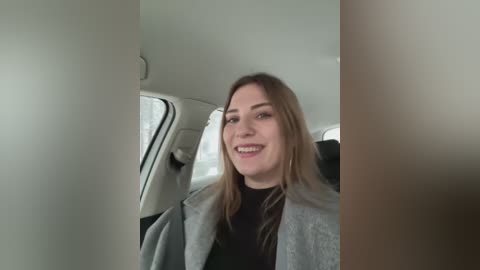 Media: Video of a smiling, light-skinned woman with long, straight brown hair, wearing a gray coat over a black top, seated in a modern car with light-colored seats.