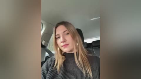 Media: Video of a young woman with long, straight blonde hair and fair skin, wearing a dark grey ribbed turtleneck sweater, sitting in a car with black interior.