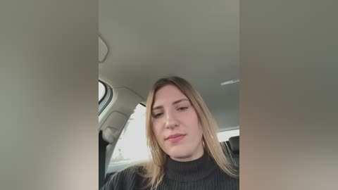 Media: Video of a young woman with straight, shoulder-length blonde hair, wearing a black turtleneck, seated in a car with beige interior, looking slightly down.