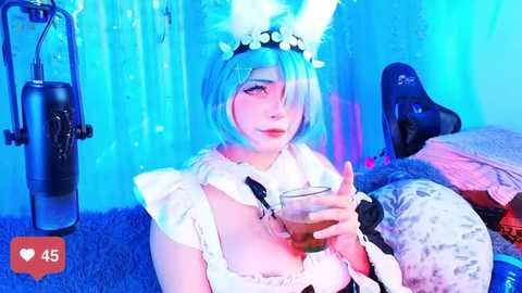 Media: A vibrant, blue-tinted video of a young woman in a cosplay bunny costume, holding a drink. She has short blue hair and wears a white frilly top. Background includes a blue curtain, gaming chair, and a furry blanket.