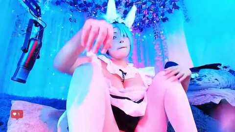 Media: Video of a young woman with blue hair and bunny ears, wearing a revealing maid outfit, sitting on a bed with a microphone, bathed in blue light.