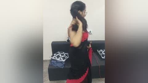 Media: Video of a woman in a black and red saree, adjusting her hair, standing next to a black sofa with patterned cushions in a simple, neutral room.