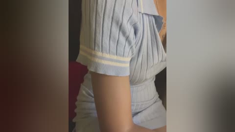 Media: Video of a person wearing a light blue, ribbed, short-sleeved polo shirt, possibly with a yellow trim. The shirt is slightly lifted, revealing a glimpse of their upper thigh. The background is blurred and dark, focusing attention on the shirt.