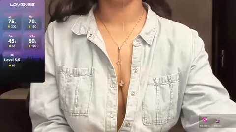 Media: Video of a person with medium brown skin, wearing a light blue denim shirt unbuttoned to reveal cleavage, adorned with a gold necklace. Background includes a virtual screen displaying health and fitness data.