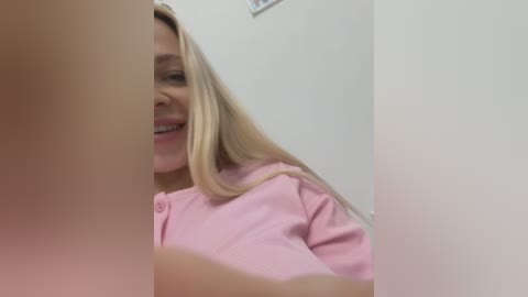 Media: Video of a smiling, blonde woman with light skin, wearing a pink button-up shirt, partially obscured by a blurry, light-colored object.