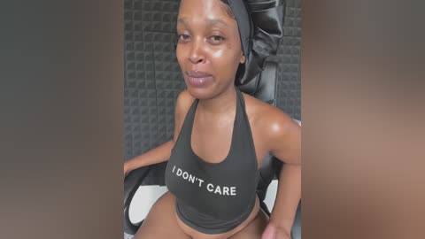 Media: A video of a dark-skinned woman with a round face, wearing a black sports bra that says \"I DON'T CARE,\" sitting in a black chair with a textured grey background.