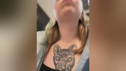 Media: Video of a blonde woman with a large, detailed black tattoo of a cat with three eyes on her chest, wearing a light blue jacket over a black top.