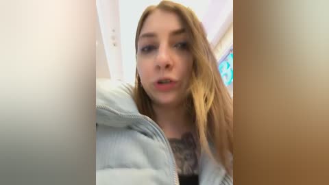 Media: Video of a young Caucasian woman with light skin, straight blonde hair, and a slightly open mouth, wearing a gray puffer jacket, taken from a low angle.