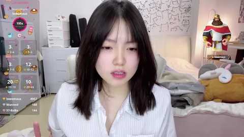 Media: Video of an Asian woman with straight black hair, wearing a white striped shirt, sitting in a cluttered bedroom with a plush toy, a lamp, and a bed visible.